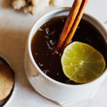 A white ceramic mug filled with a dark liquid garnished with a cinnamon stick and a slice of lime—one of those must-try drinks. Next to the mug, a small bowl holds several round cookies. The background surface is light-colored and textured.