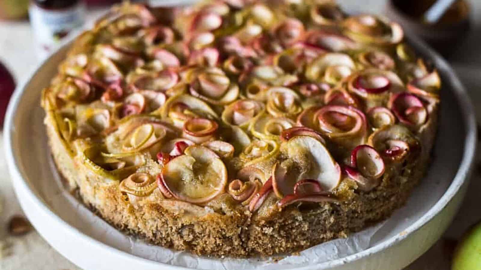 A close-up of a round apple tart on a white plate. This forgotten dessert classic features thinly sliced apples arranged in a decorative, overlapping pattern on top of a crumbly crust. The apples have red and yellow skins, giving the tart a colorful appearance.