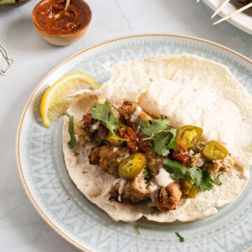 A plate of tacos with grilled chicken, topped with cilantro, jalapeños, and a sprinkle of lemon, accompanied by sauces on a light-colored table.