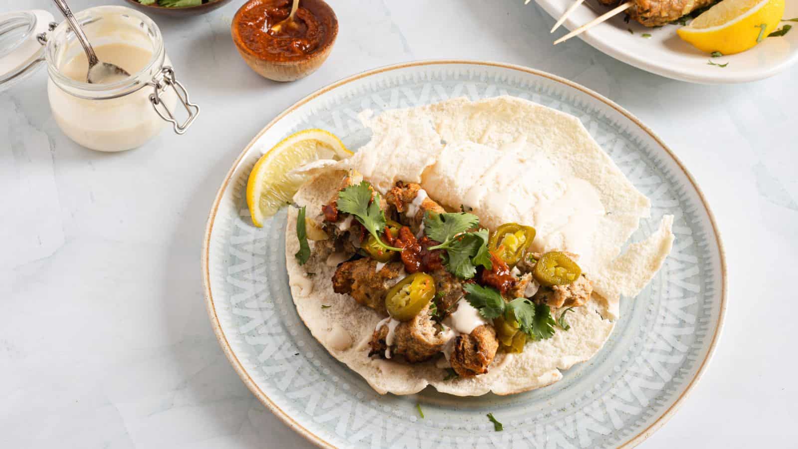 A plate of tacos with grilled chicken, topped with cilantro, jalapeños, and a sprinkle of lemon, accompanied by sauces on a light-colored table.