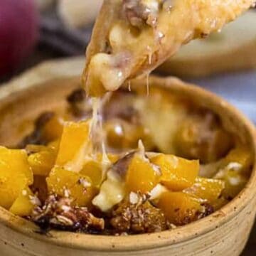 A piece of bread being dipped into a 30-minute baked cheese dish with nuts and fruit pieces.