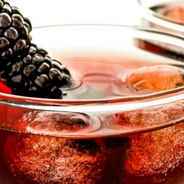 Two glasses of red cocktail with ice cubes, garnished with a blackberry on top, against a white background.