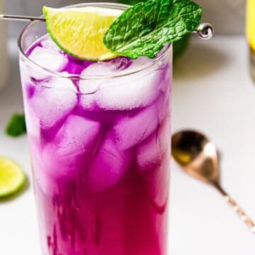 A vibrant purple beverage with ice in a tall glass, garnished with a lime wedge and a mint leaf, beside a bottle and slices of lime on a white surface.