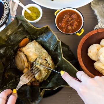 A person is unwrapping a traditional dish cooked in banana leaves, featuring Grandma's Chicken Recipes, with side dishes and condiments placed around on a table.