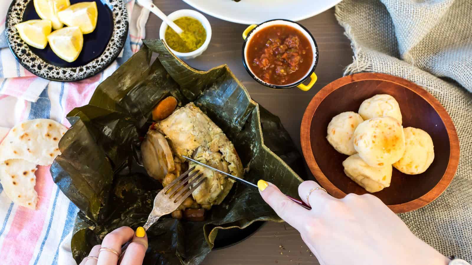 A person is unwrapping a traditional dish cooked in banana leaves, featuring Grandma's Chicken Recipes, with side dishes and condiments placed around on a table.
