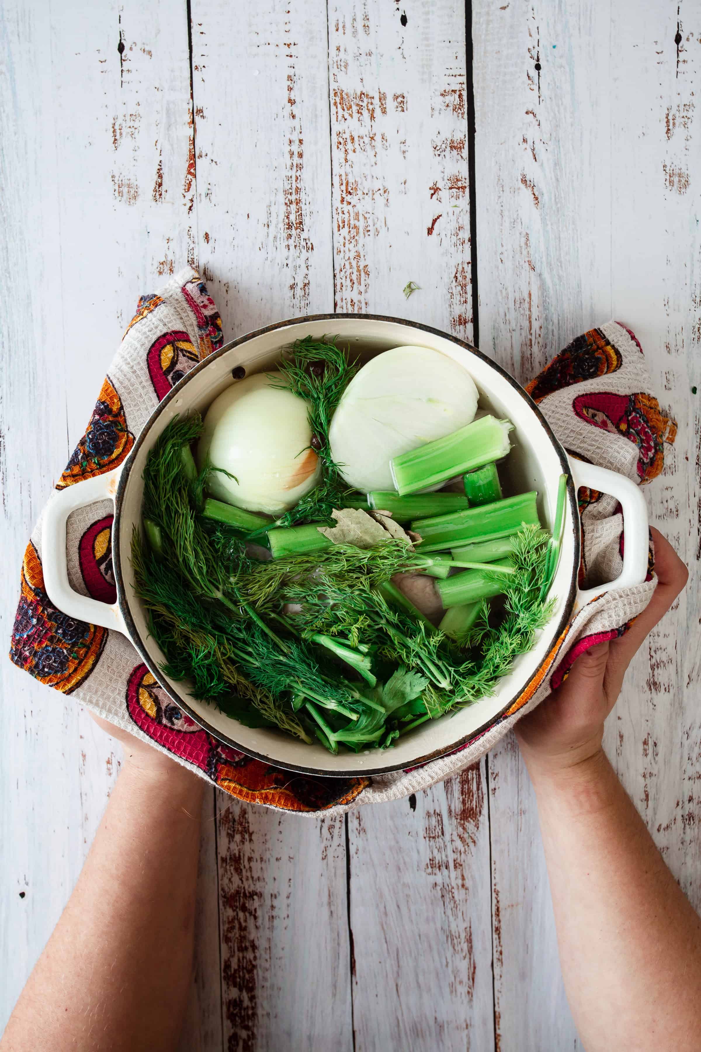 Hands holding a pot filled with fresh fennel, dill, and halved onions on a rustic wooden surface.