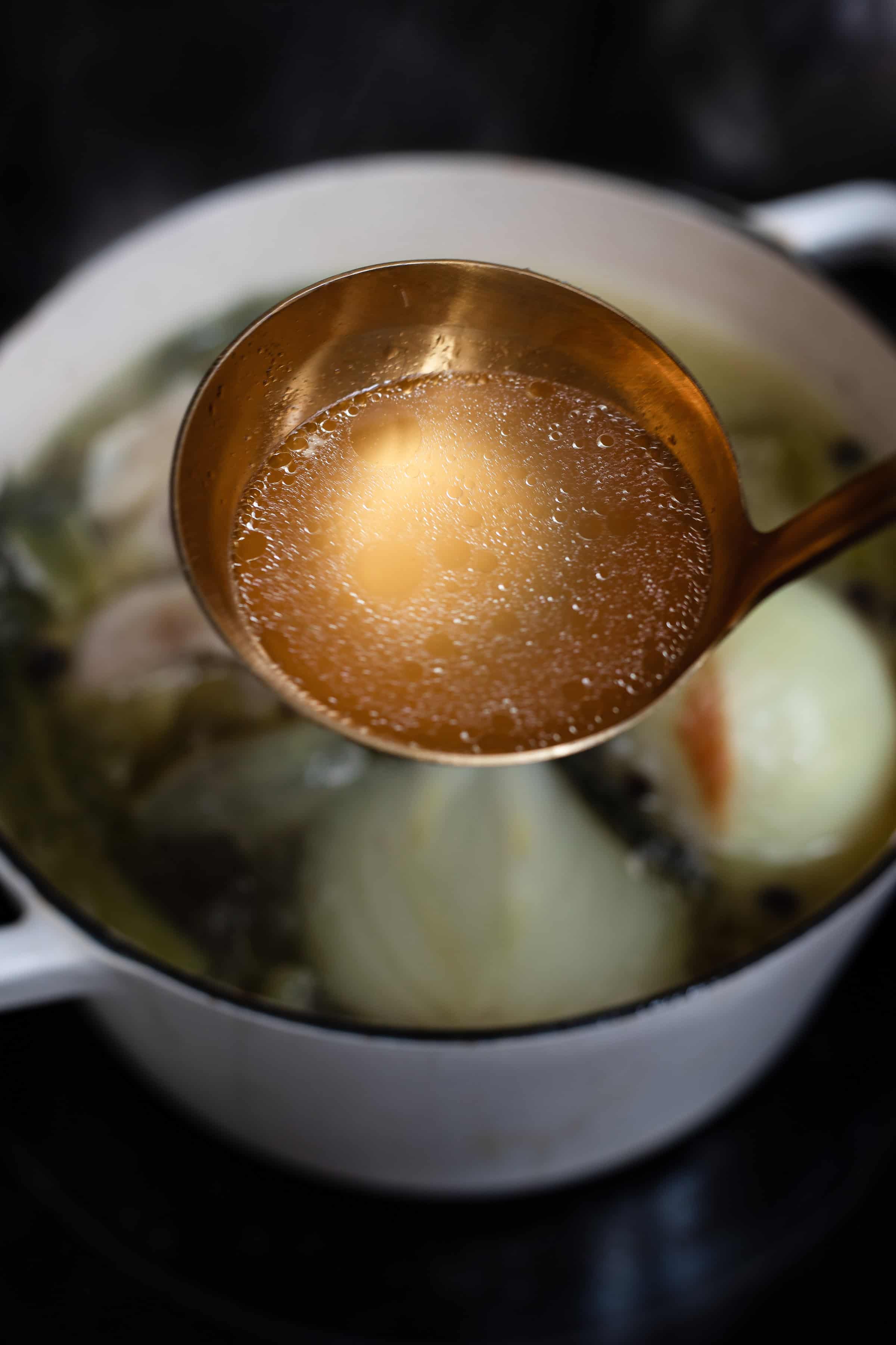 A ladle is being used to scoop broth from a pot containing onions and herbs, demonstrating the preparation of a soup.