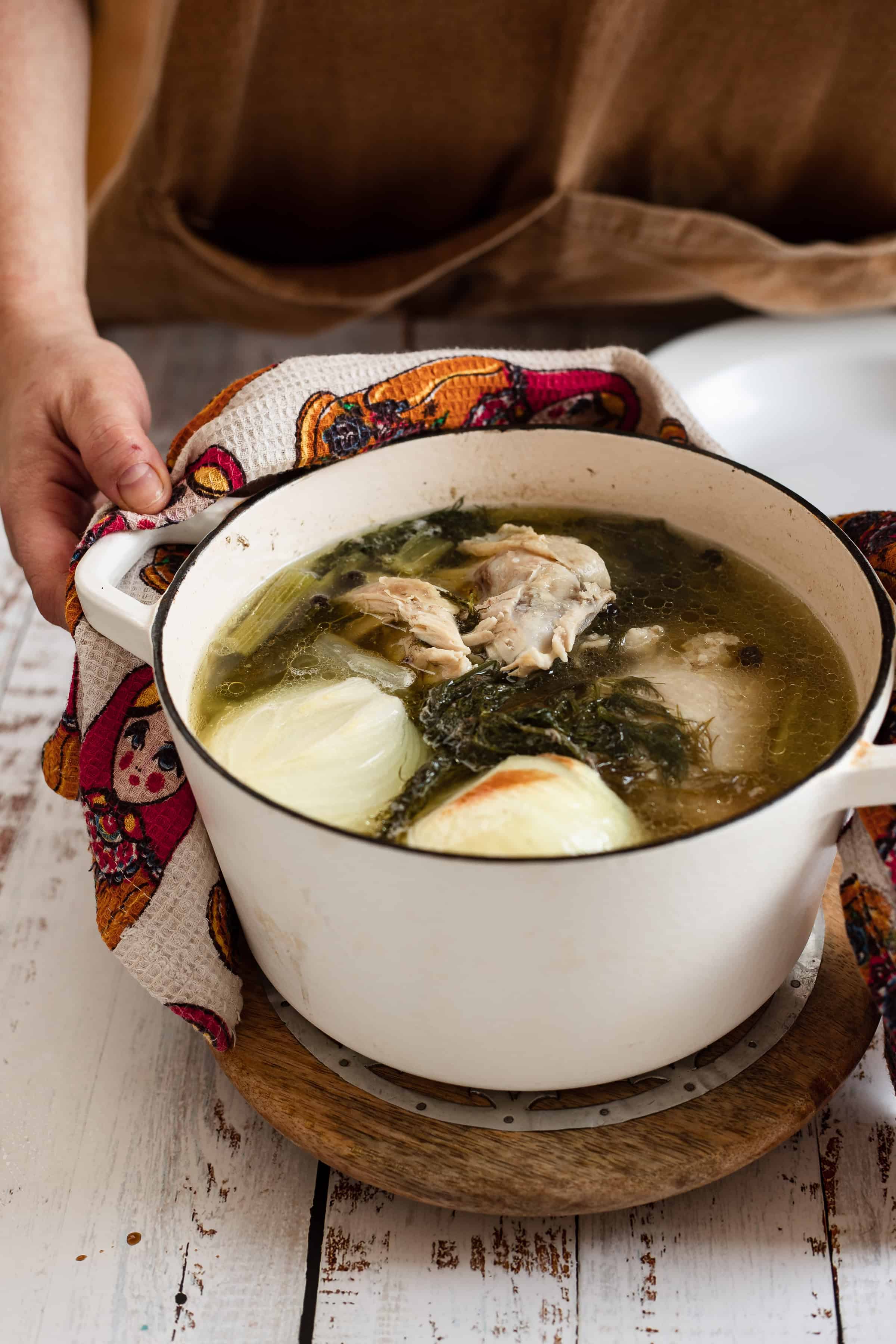 A person's hand holds a colorful cloth next to a white pot filled with chicken soup, featuring onions, greens, and visible broth, on a wooden trivet.