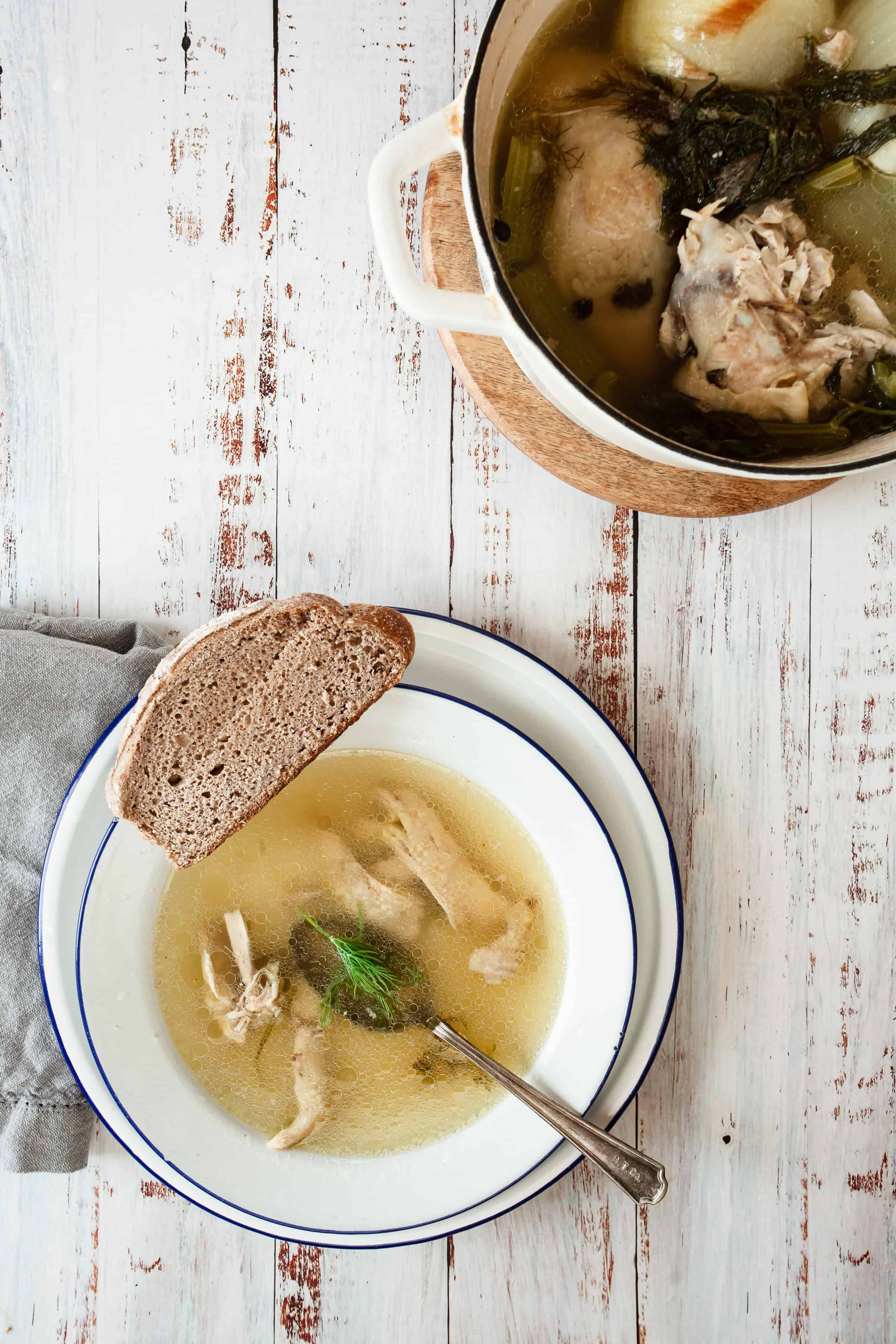 A bowl of chicken soup with a piece of bread on a rustic wooden table. The soup contains pieces of chicken and is served in a white bowl with a blue rim alongside an auto draft metal spoon.