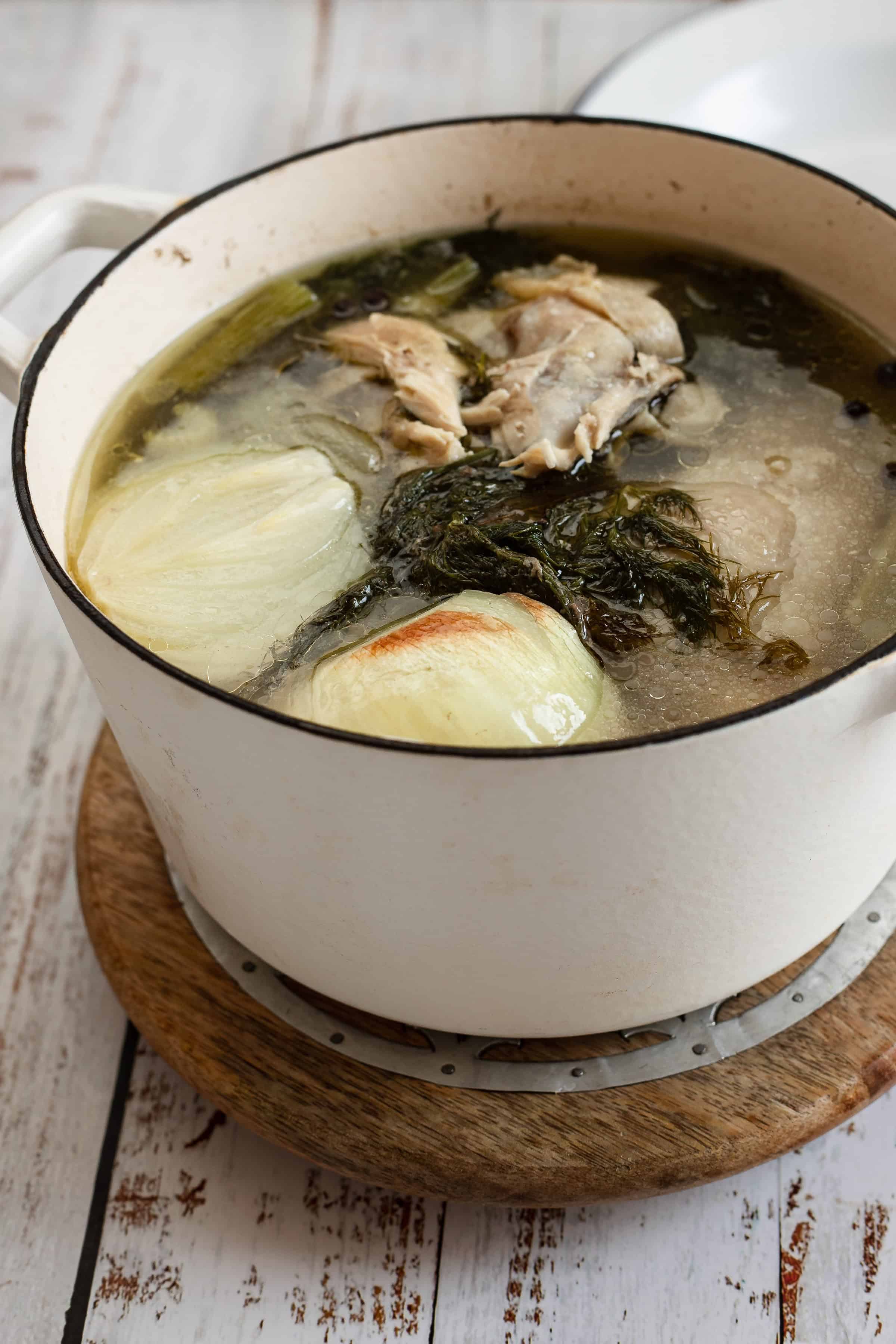 A pot of chicken soup on a wooden table, featuring whole chicken pieces, onions, and leafy greens simmering in a clear broth.