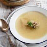 A bowl of Jewish chicken soup with a sprig of dill and pieces of chicken, served in a white bowl with blue trim on a wooden table, accompanied by a wooden spoon.