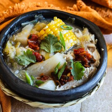 A traditional colombian soup called ajiaco served in a black bowl, garnished with corn, capers, and fresh herbs, accompanied by a glass of dark-colored beverage on a textured tabletop.