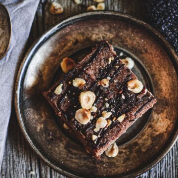 A single square of chocolate brownie garnished with chopped nuts is placed on a rustic metal plate. A vintage spoon rests on a folded gray cloth to the left, and a dark textured fabric is seen on the right. The background is a weathered wooden surface, evoking the charm of forgotten dessert classics.