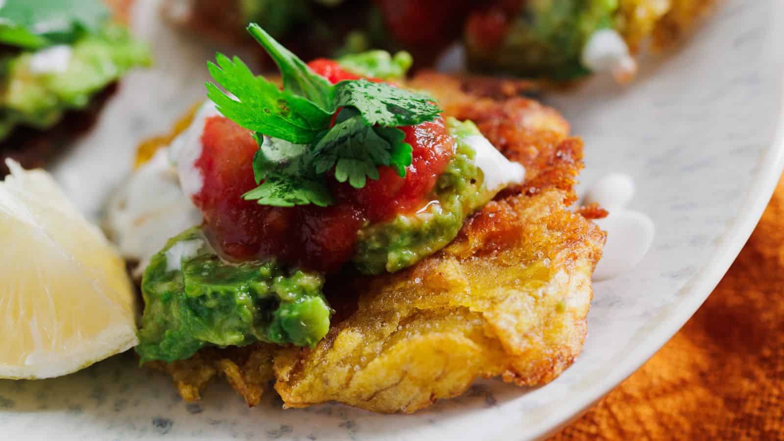 A close-up of a plated dish featuring crispy plantains topped with guacamole, salsa, a dollop of sour cream, and garnished with fresh cilantro. A lemon wedge is visible to the side of the dish. The plate rests on an orange tablecloth, evoking treasured family recipes shared around the dinner table.