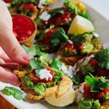 A hand holding a tostone topped with guacamole, salsa, and a cilantro leaf. The plate is filled with similar tostones, with lemon wedges and a small bowl of salsa in the background. Perfect for adding to your weekly menu, these delicious bites are showcased on a white rectangular plate resting on an orange cloth.
