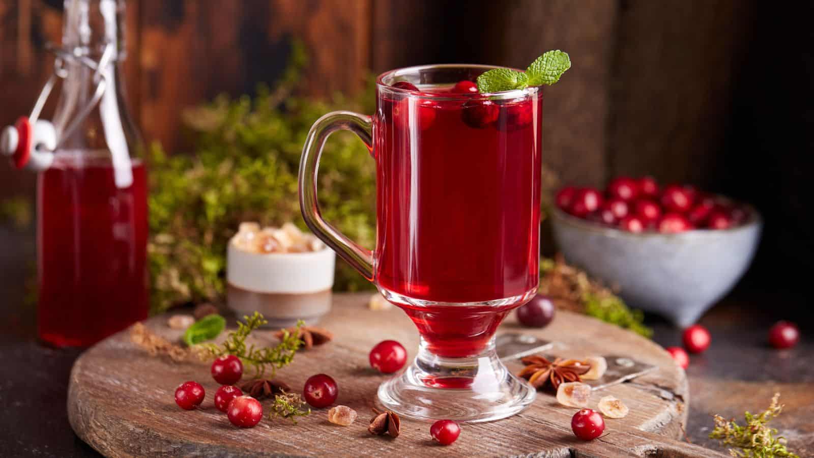 A glass mug filled with hot cranberry drink garnished with mint on a wooden surface, surrounded by cranberries, star anise, and a candle, with a bottle and bowl of cranberries in