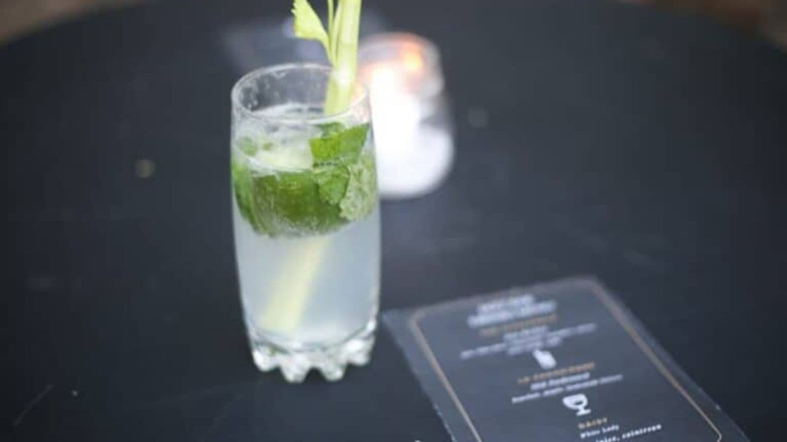 A highball glass containing a light green cocktail with ice cubes, mint leaves, and a celery stalk is placed on a dark surface. A menu card of must-try drinks lies beside the glass, and a lit candle is visible in the background.