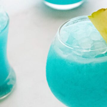 Two vibrant blue cocktails served over ice in round glasses, garnished with a slice of pineapple on the rim of one glass, presented on a white surface.