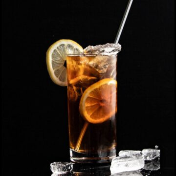 A glass of iced tea with lemon slices, garnished with a lemon wedge on the rim and a stirring straw, surrounded by scattered ice cubes on a dark background.