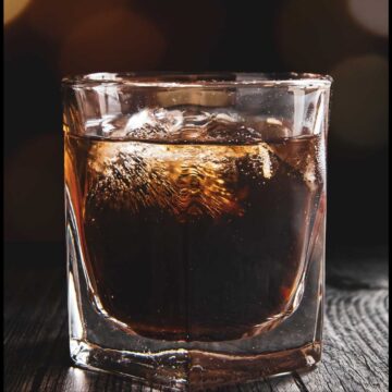 A glass of whiskey with ice on a wooden surface, illuminated by soft background lighting that creates a warm, inviting glow, reminiscent of drinks at grandma's house.