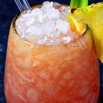 Close-up of a chilled cocktail in a textured glass, topped with crushed ice, garnished with a slice of pineapple and a green straw, on a dark textured surface.
