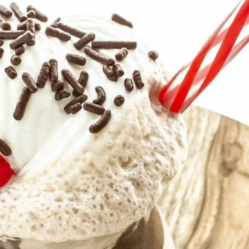 A close-up image of a vanilla milkshake topped with whipped cream, chocolate sprinkles, and a red cherry, served in a glass with a red and white striped straw.