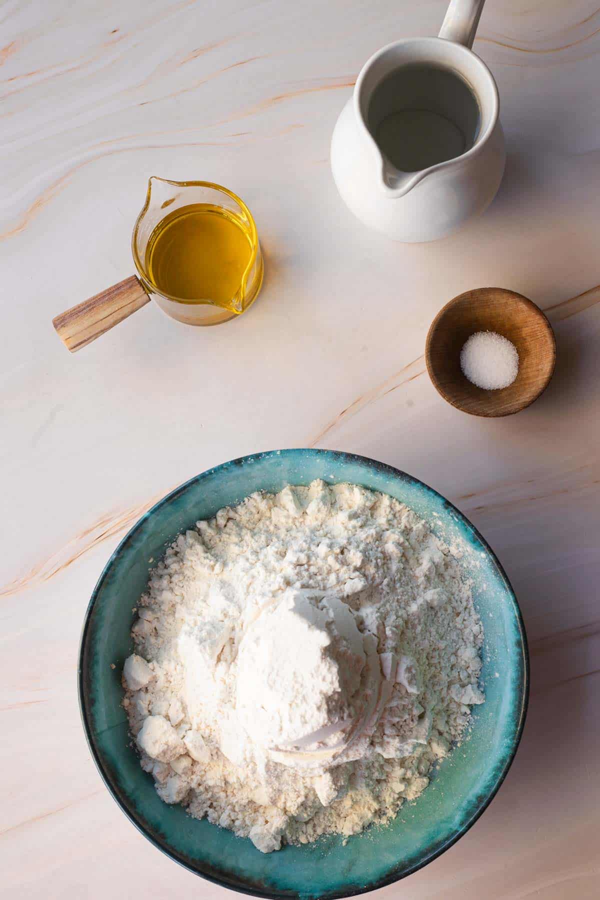 A top-down view of baking ingredients on a table for a gluten-free matzo recipe, featuring a bowl of flour, a jug of milk, a cup of oil, and a small bowl of salt