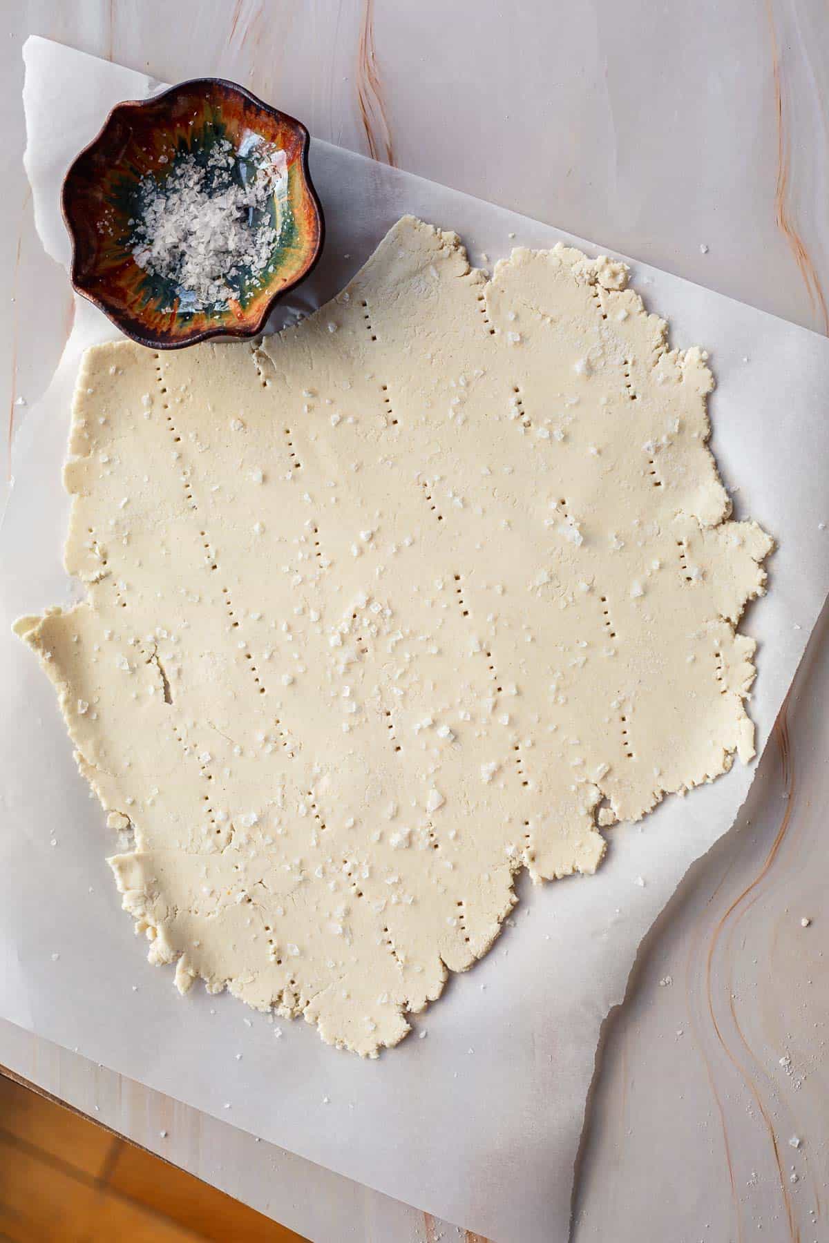 A batch of gluten-free matzo dough rolled out on parchment paper beside a small, colorful bowl filled with flour. The surface of the dough is textured with small indentations.