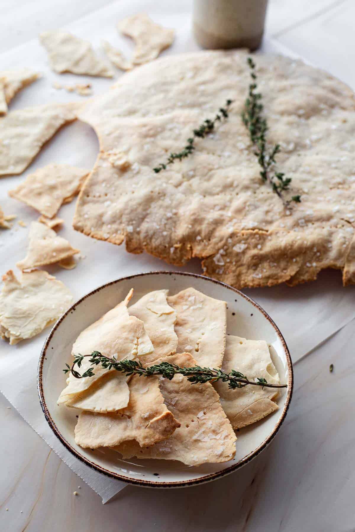 A plate of freshly baked gluten-free matzo with thyme, surrounded by scattered pieces and a rolling pin, on a wooden table.