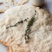 A freshly baked gluten-free flatbread with a sprig of thyme on top, accompanied by a ceramic jug and scattered crumbs, presented on a light surface.