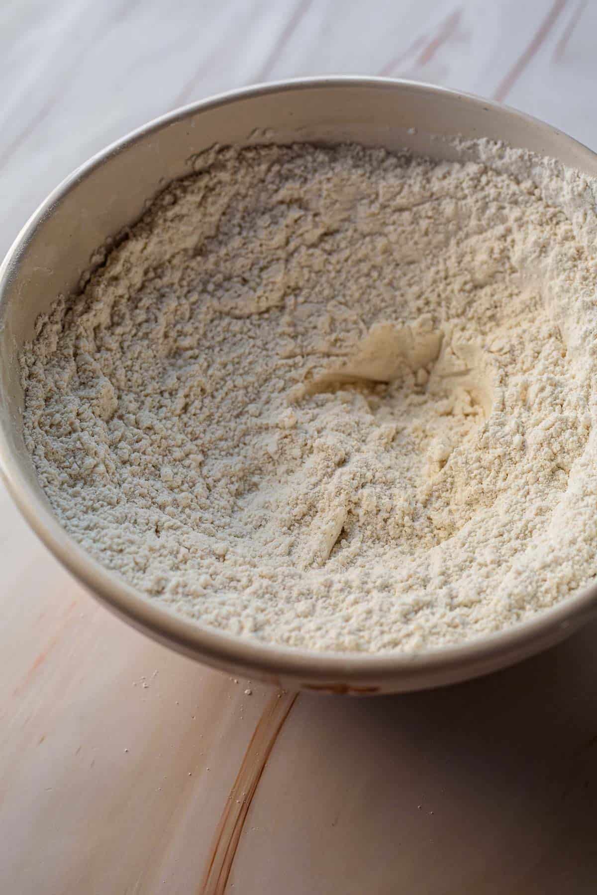 A close-up image of a bowl filled with gluten-free flour, placed on a light wooden surface. The texture of the flour is finely ground and loosely settled.
