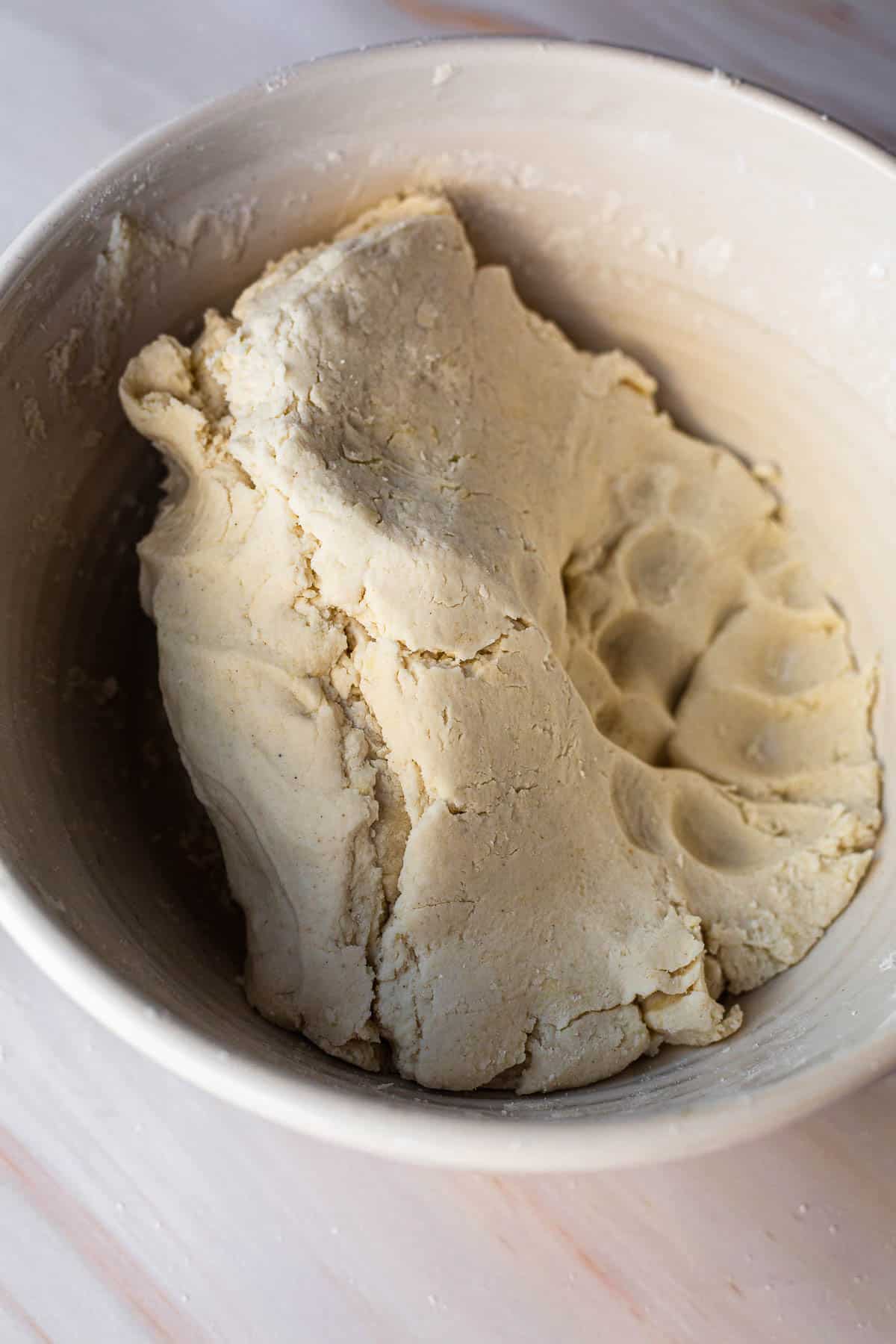 A close-up image of a bowl containing a freshly prepared, slightly textured gluten-free matzo dough, with visible indentations and creases, set on a light wooden surface.