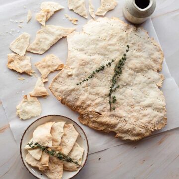 Large, thin homemade cracker with salt and herbs, partially broken, on a wooden surface with some pieces on a small plate and a sprig of thyme on top. In the background sits a