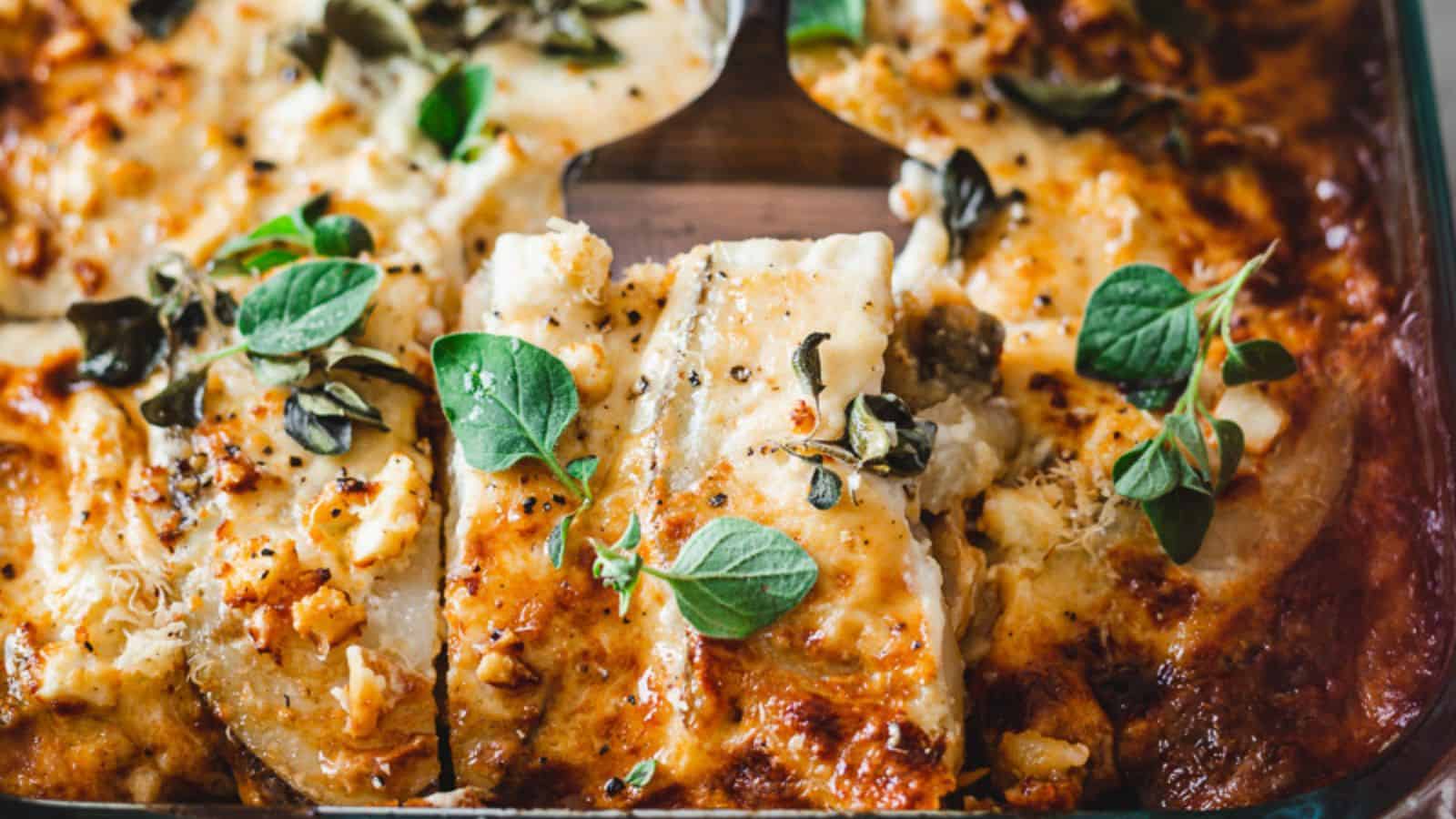 A close-up image of a baked casserole with layers of sliced potato topped with melted cheese, garnished with herbs and capers in a glass dish.