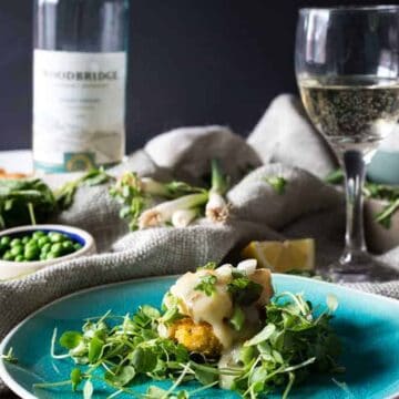 A meal is served on a blue plate with a garnish of greens and a creamy sauce. In the background, there are fresh vegetables, a bowl of peas, a white sauce bottle, and a glass of white wine. The setting is decorated with a light-colored cloth, perfect for those who love quick recipes.