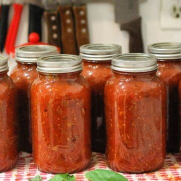 Eight jars of tomato sauce with metal lids are arranged in two rows on a countertop, promising some of the best things you'll eat. The jars rest on a red and white striped towel. In the background, kitchen knives are visible in a knife block, ready for culinary magic.