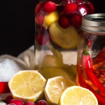 Two jars of must-try drinks are displayed against a dark background. One jar contains lemon slices and cranberries, while the other has red chili peppers and herbs. In front of the jars are halved lemons, cranberries, and a whole red chili pepper.