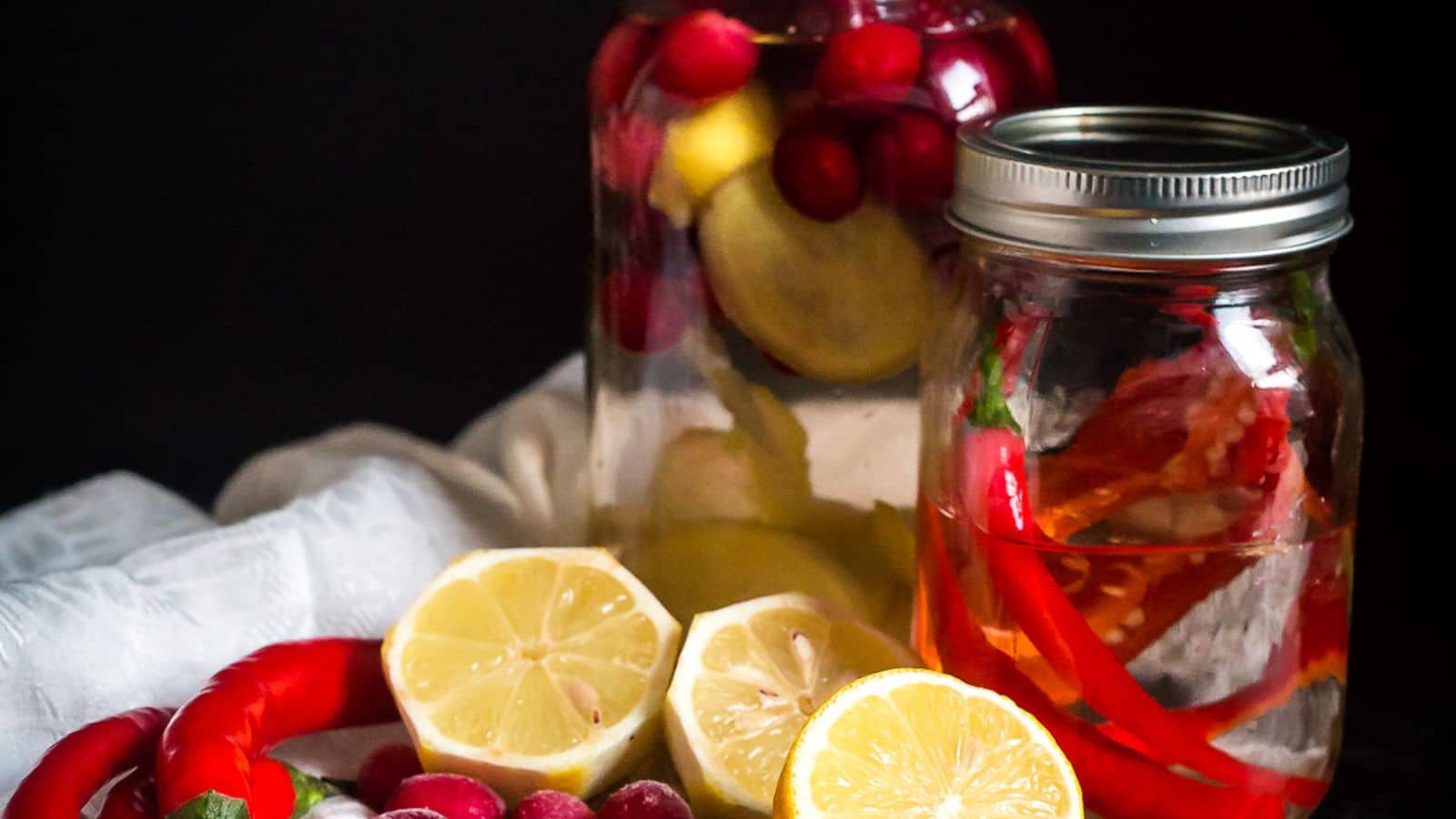 Two jars of must-try drinks are displayed against a dark background. One jar contains lemon slices and cranberries, while the other has red chili peppers and herbs. In front of the jars are halved lemons, cranberries, and a whole red chili pepper.