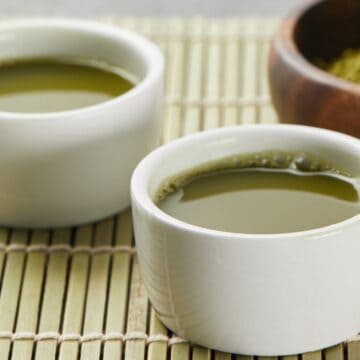 Two white ceramic cups filled with green tea sit on a bamboo mat. A wooden bowl containing green powder is visible in the background, partially out of focus, showcasing one of the must-try drinks for any tea enthusiast.