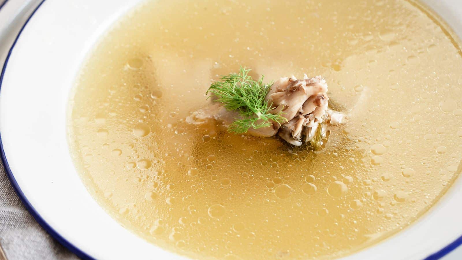 A bowl of Jewish chicken soup with a piece of chicken and dill on top, served in a white bowl with a blue rim. The soup appears hot with visible small bubbles on the surface.