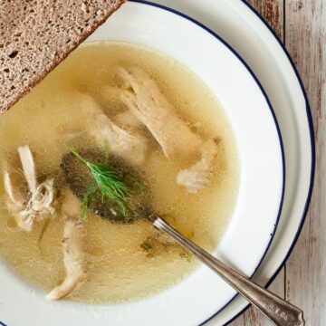 A white bowl filled with clear Jewish chicken soup, featuring pieces of chicken, fresh dill, and a silver spoon. The bowl is placed on a white wooden table with a slice of brown bread resting on its edge. A grey cloth napkin is partially visible next to the bowl.