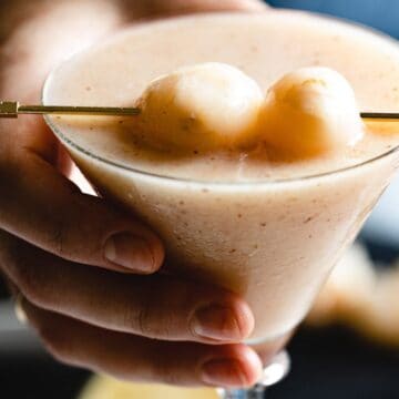 A hand holds a frosted cocktail glass filled with a light-colored drink. Two lychees are skewered on a stick and placed on top of the glass as garnish. The background is slightly blurred with indistinct objects, evoking the elegance often found in sophisticated drink recipes.