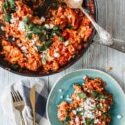 A skillet with tomato and scrambled egg mixture, known as Chilaquiles, garnished with herbs and white cheese, next to a plate serving the same dish, accompanied by utensils and a cloth