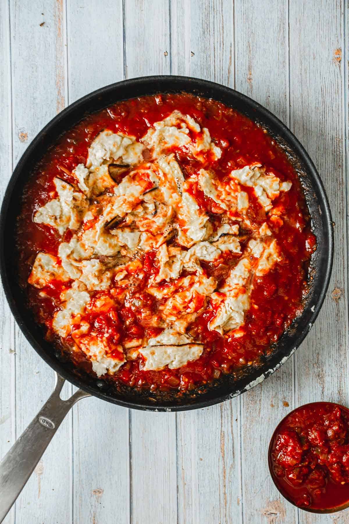 A skillet with tomato sauce, chunks of white cheese on a wooden surface, featuring Mexican Matzo Brei.