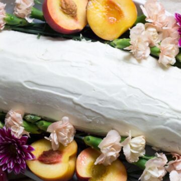 A white-frosted cake decorated with pink and purple flowers and fresh peach slices on a light-colored surface, inspired by grandma's secret recipes.