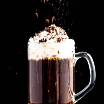 A glass mug of hot chocolate topped with whipped cream and chocolate shavings, with more shavings sprinkling down, set against a dark background.