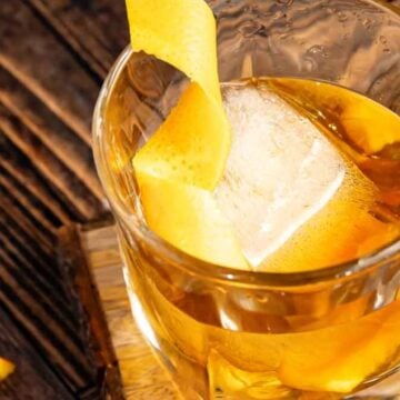 A glass of whiskey with a large ice cube and a twist of lemon peel, standing on a wooden board beside slices of orange.