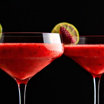 Two glasses of red fruit smoothie garnished with lime slices and raspberries against a black background.