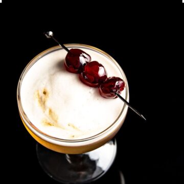 A cocktail with a creamy, frothy top garnished with three cherries on a skewer, set against a stark black background, highlighting the contrast and rich colors of the drink.