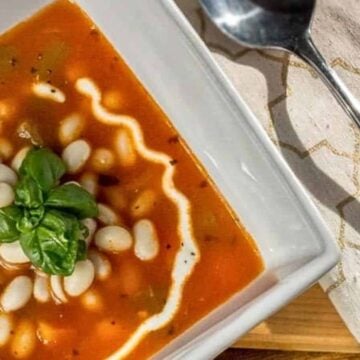 A bowl of tomato-based soup garnished with white beans and a basil leaf, with a swirl of cream on top. It is served in a rectangular white dish with a metal spoon and a patterned beige napkin placed beside it—perfect for family recipes to try this week.